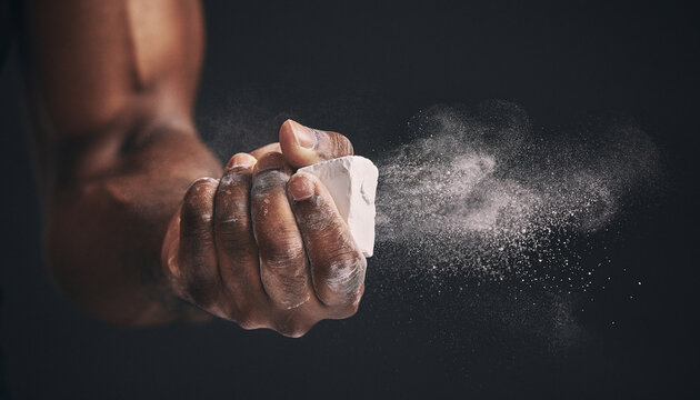 Close-up of an Olympic athlete's hand crushing a chalk block, creating a dramatic powder explosion against a blurred gym background with heavy weightlifting equipment.
