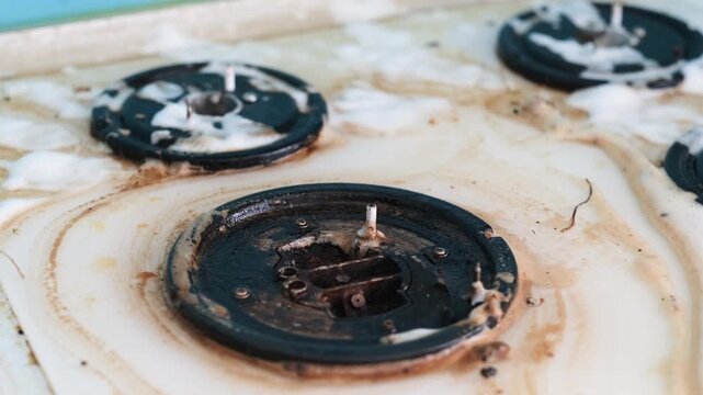 A woman's hand in a rubber glove splashes foam and washes a dirty gas stove.

