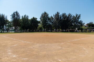 快晴の奥に木々のある赤土のグラウンド