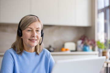 Naklejka premium Senior woman with headset smiling during a laptop video call from her home kitchen, working remotely as a confident, tech-savvy consultant connecting with clients worldwide