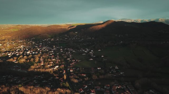 Aerial view of Ascain with houses nestled amongst the trees, bathed in golden sunlight contrasting with deep shadows, creating a picturesque scene, Ascain, Nouvelle-Aquitaine, France.