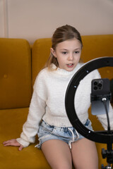 Girl sitting on yellow sofa and filming video content using smartphone and ring light, representing...