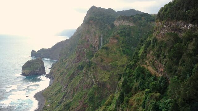 Aerial view of Cascata da Rocha do Navio waterfall dropping down steep cliffs in Madeira, drone descending on a moody morning with bright light breaking through clouds over the Atlantic ocean.