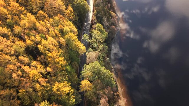 Aerial view of a winding road running alongside a dark, reflective body of water, framed by a forest with autumnal hues, Callander, Scotland, United Kingdom.
