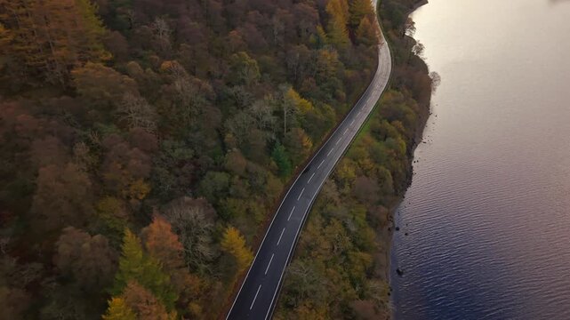 Aerial view of a road winding along the edge of Loch Lubnaig, contrasting with the dark blue water and the colorful autumn foliage, Callander, Scotland, United Kingdom.