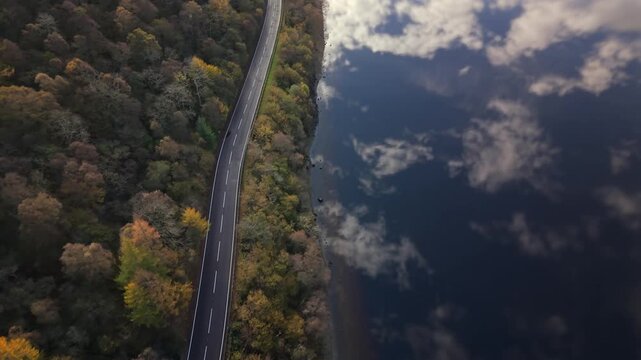 Aerial view of a winding road running alongside a lake reflecting the sky, contrasted with the autumn colors of the adjacent trees, Callander, Scotland, United Kingdom.