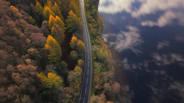 Aerial view of a road running between Loch Lubnaig and a forest in autumn colors, reflecting the sky in the water, Callander, Scotland, United Kingdom.