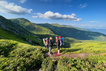 Obraz premium Two hikers, man and woman with backpacks and sleeping pads, stand facing each other on mountain trail. Stunning green hills under bright blue sky on background.