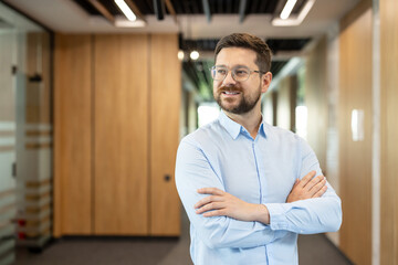 Confident businessman in glasses and a light blue shirt stands with arms crossed, smiling in a bright modern office, conveying leadership, success and professional growth © Liubomir