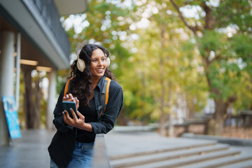Fototapeta premium Young Hispanic college student with headphones smiling while listening to music on her smartphone outdoors on campus, casual backpack, enjoying a study break in autumn green surroundings