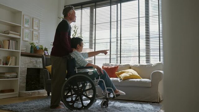 Happy senior Asian husband caring for his disabled wife in wheelchair at home.Concept of eternal love,elderly healthcare insurance,Relationship,trust and supportive retirement life for aged people.