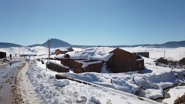 HD moving shot pushing forward into a snow-covered rural village in the Middle Atlas. Features traditional houses, a winter road, and snowy mountain landscapes under a blue sky.
