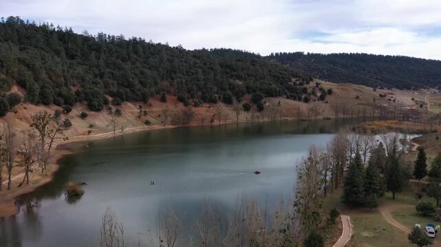 wide shot of a tranquil mountain lake surrounded by cedar forests. Features calm water reflections and seasonal trees along the shoreline in the Middle Atlas.