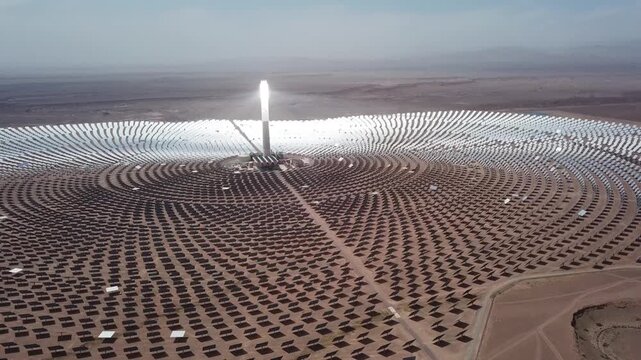 Aerial drone shot of the Noor III Concentrated Solar Power (CSP) plant. Features the glowing central tower surrounded by thousands of heliostat mirrors in the Moroccan desert.