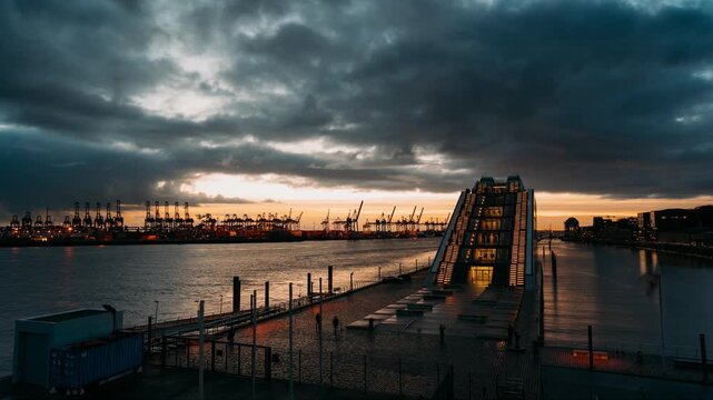 Passing clouds and ships during sunset in Hamburg harbour and container terminal on the river Elbe with illuminated lights on a modern architecture glass building