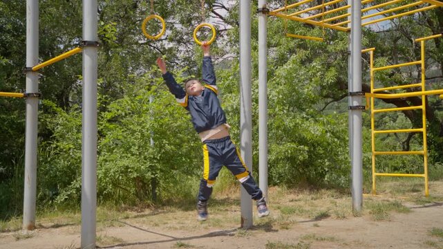 Active child training on yellow gymnastic rings against green trees background. Outdoor workout, developing strength and coordination during summer day.