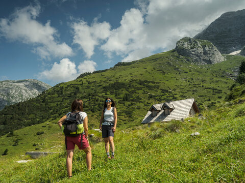 Two hikers stand on a grassy hillside with a mountain hut,  Casera Canin, Natural Julian Alps Park, and peaks in the background under a partly cloudy sky. Casera Canin,Resia Valley,Julian Alps,Italy