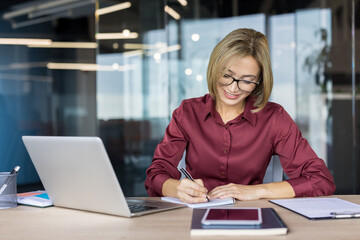 Fototapeta premium Confident businesswoman with eyeglasses smiling at her modern desk, writing notes in a spiral notebook with laptop and smartphone nearby, organized and focused on work
