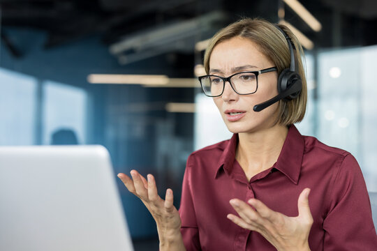 Stressed customer service representative wearing a headset, communicating with clients and indicating confusion or problems while working on a laptop in a modern office