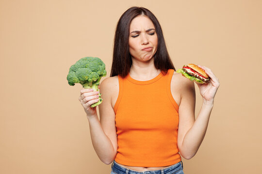 Young minded woman wear orange casual clothes hold eat choose between broccoli and burger isolated on plain pastel light beige background. Proper nutrition healthy fast food unhealthy choice concept.