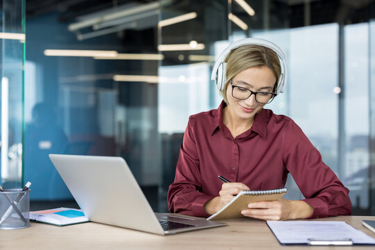 Young woman wearing headphones and glasses, watching webinar on laptop, writing notes in a notebook, studying or working remotely in a modern office setup
