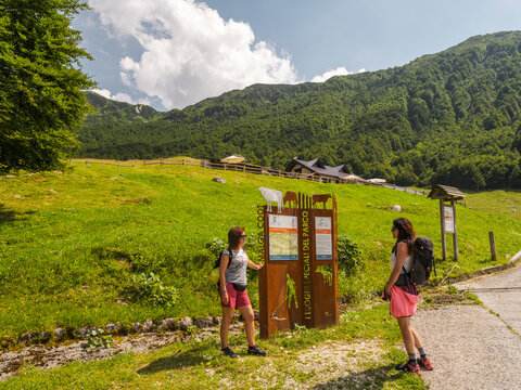 Two women hikers look at a sign in a grassy meadow with mountains in the background. Casera Coot,Resia Valley,Julian Alps,Italy