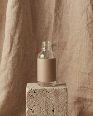 Naklejka premium A clear bottle with a blank beige label stands on a porous stone cube. The background consists of draped, textured beige fabric, creating a monochromatic and minimalist aesthetic.