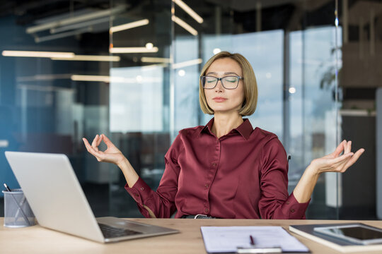 Young businesswoman with eyes closed and hands in mudra gesture, finding inner calm and mental well-being while taking a mindful break from work at her office desk