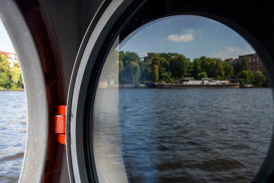 Boat porthole window on the Spree river in Berlin Germany framing water and shoreline trees in summer with a calm travel view