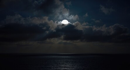 Bright full moon glowing through dark clouds over the sea at night