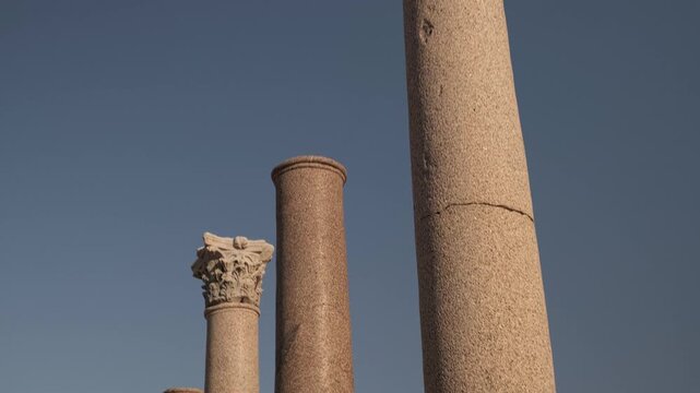 Tilt up footage of tall ancient stone columns rising in foreground, with modern residential buildings visible behind them. Kom el-Dikka archaeological site in Alexandria, Egypt