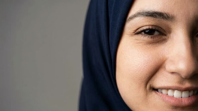 Half-face close-up of a happy Middle Eastern woman in a blue hijab smiling at the camera. Portrait of a cheerful Muslim lady showing white teeth and positive emotion. Islamic culture and beauty 