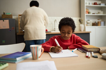 Naklejka premium Black child drawing with marker at table, focused on artwork, while middle aged African American woman standing in kitchen background, books and mug placed nearby on surface