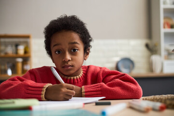 Fototapeta premium Portrait of Black child sitting at desk writing in notebook, looking directly at camera with serious expression, hands holding pen, background showing blurred kitchen interior
