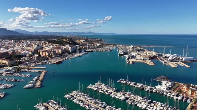 aerial view marina with yachts, turquoise water, sunlit docks, crowded berths, coastal town backdrop, mountain
