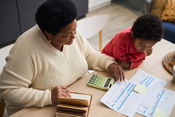 Middle aged Black woman reviewing financial documents with calculator while young Black child...