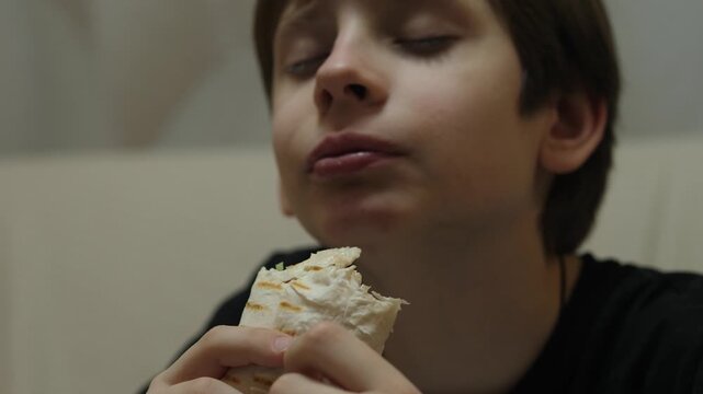 teenage boy eating shawarma with appetite in a home setting. The child enjoys the taste of fast food, squinting his eyes with pleasure during a snack
