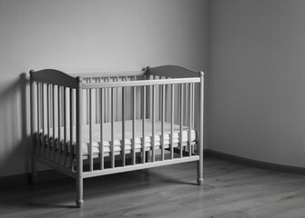 A simple, empty wooden baby crib with a mattress stands in the corner of a dimly lit, minimalist room.