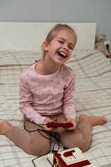 Cheerful girl generation Alpha playing a Millennial retro game console while sitting on the bed. Winning a video game. Nostalgia for the 80s and 90s. Vertical orientation.