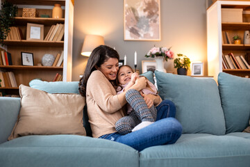 Obraz premium Happy mother tickling her little daughter on the sofa. Family fun, bonding and motherhood concept. Cheerful woman playing with laughing child in a cozy living room during leisure time at home.
