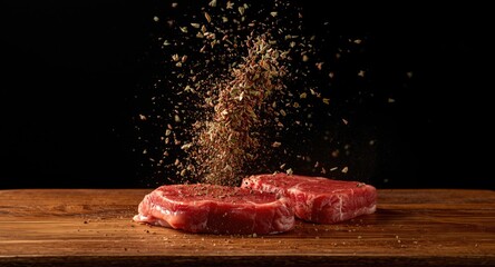 Action shot of herbs and spices in flight landing on raw beef steaks over wooden surface black background