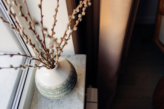 Pussy willow branches in a ceramic vase on a windowsill with warm natural light. Cozy minimalist home decor, spring atmosphere, neutral tones and soft focus.