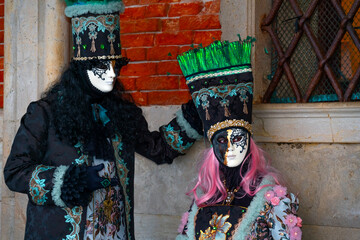 Portrait of Venetian carnival couple wearing matching historical costumes and elegant masks, standing together against the textured architecture of Venice