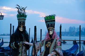 Elegant couple in traditional Venetian carnival masks and historical costumes posing by gondolas in Venice. Concept of mystery, luxury travel and European tradition