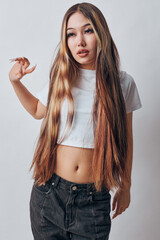 Young woman with long straight hair wearing white crop top and black pants posing on white background with hand raised and thoughtful expression in studio shot.