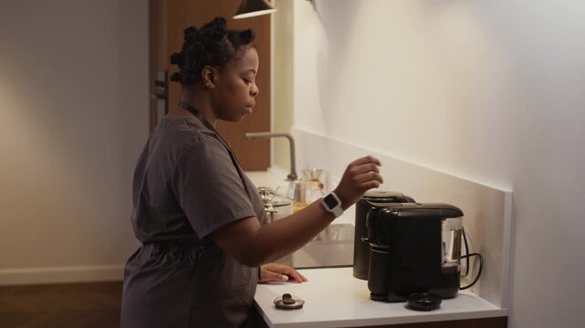 African American woman dressed in scrubs making coffee with help of mini coffee machine while having break at home