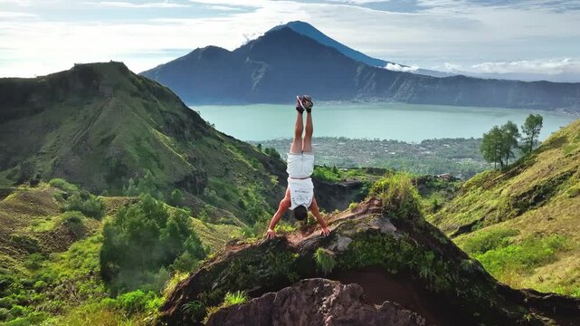 Young man doing handstand on mountain ridge, embracing freedom and challenging personal limits while traveling in Bali with stunning volcanic landscape and lake Batur in the background. Aerial flight