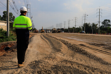Civil engineer in reflective safety vest and helmet monitoring road construction site, coordinating machinery, traffic control, and utility infrastructure along expanding highway corridor. © DK_STUDIO