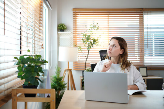 caucasian woman in home office, using laptop for research in remote work, ideas and thinking. Happy girl at desk with computer, writing notes and online search in house for freelance networking
