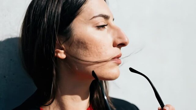 female big nose - A woman with closed eyes and sunglasses hanging from her mouth, leaning against a textured wall, exudes calmness and contemplation in a minimalist setting